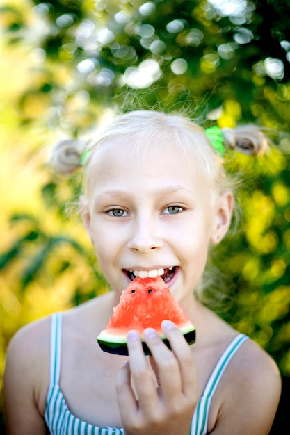 Adorable blonde girl eats a watermelon outdoors