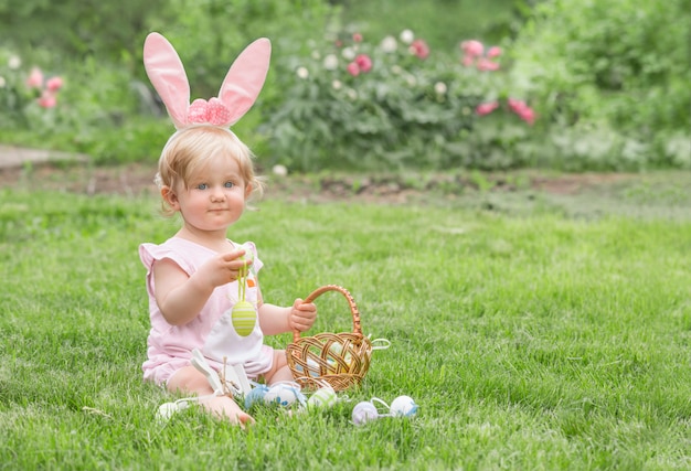 Adorable blonde toddler girl wearing bunny ears playing with Easter eggs in a basket sitting in a sunny garden