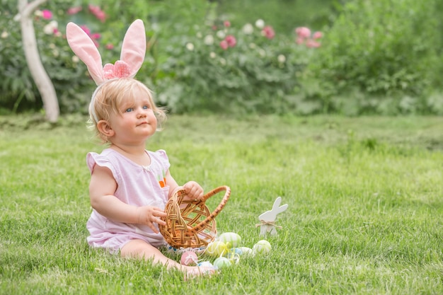 Adorable blonde toddler girl wearing bunny ears playing with Easter eggs in a basket sitting in a sunny garden