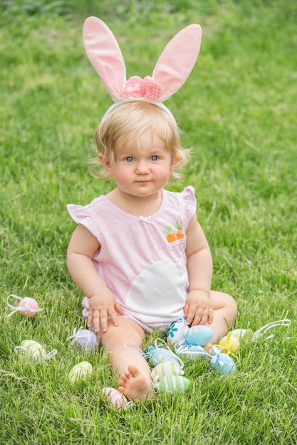 Adorable blonde toddler girl wearing bunny ears playing with Easter eggs in a basket sitting in a sunny garden