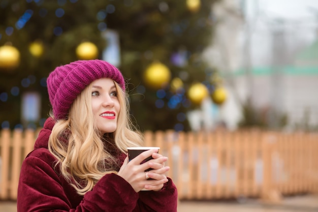 Adorabile donna bionda vestita con abiti invernali e che beve caffè all'albero di natale