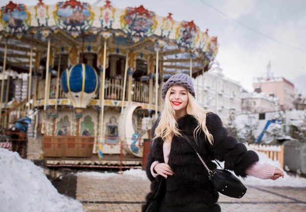 Adorable blonde woman wearing knitted hat and fur coat, posing near the carousel