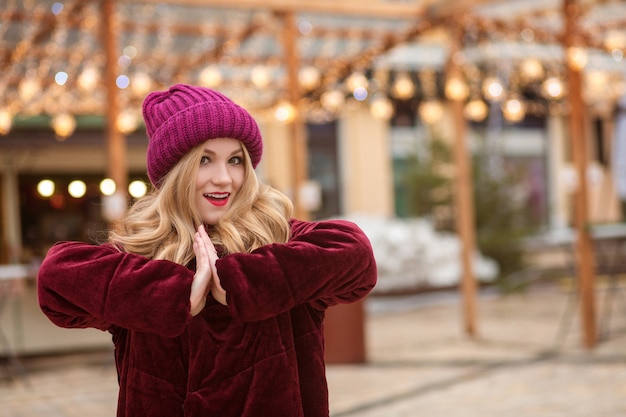 Adorable  blonde woman wearing red knitted hat and winter coat, posing on the background of garland at the street in Kyiv