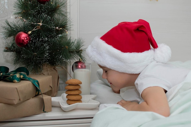 愛らしい男の子は、サンタクロース、空きスペースのためにミルクとクッキーを調理します。伝統的な御馳走は、ベッドの近くのベッドサイドテーブルにクリスマスクッキーとミルクのグラスです。新年のコンセプト。親族