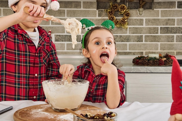 Adorable boy kneads dough with a wooden spoon when his cute\
younger sister climbs her hands into the dough and tastes it.\
charming caucasian children cooking christmas cookies together at\
home kitchen