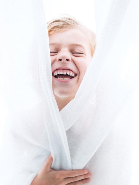 Adorable boy laughing with closed eyes and mouth opened covering body with white curtain