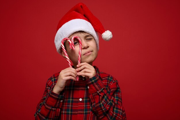 산타 모자와 격자 무늬 셔츠를 입은 사랑스러운 소년은 하트 모양의 사탕 사탕 지팡이를 들고 크리스마스 광고를 위한 복사 공간이 있는 빨간색 배경에 포즈를 취한 카메라를 들여다봅니다.