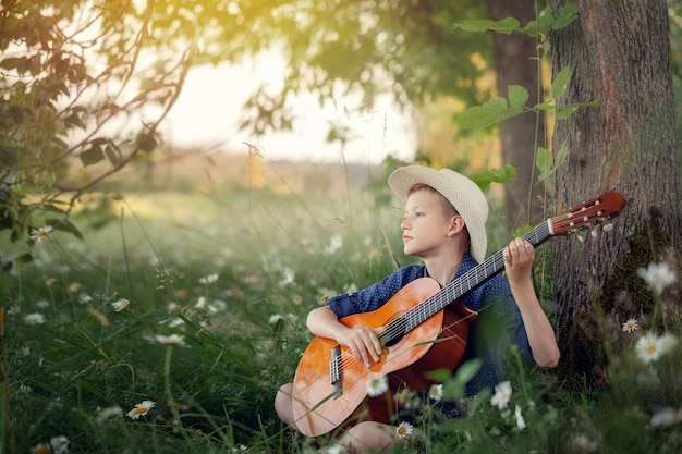 Adorable boy with guitar, relaxing in the park. Kid sitting on a grass in summer day