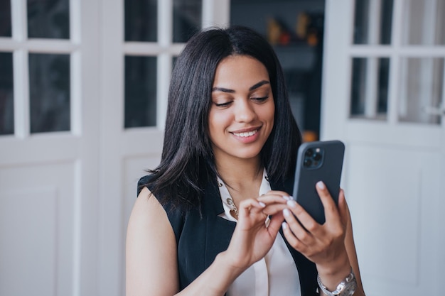 Adorable Brazilian girl holding phone typing text message to her boyfriend smiling