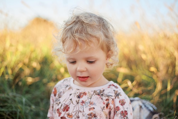 Adorable Caucasian blonde wavy haired baby girl outdoors at sunny summer day looking away walking playing alone in field not far from house on beautiful summer sunset