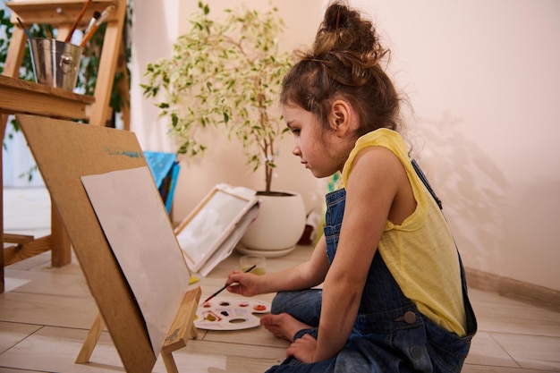 Adorable Caucasian child cute little girl sitting on the floor at a wooden easel and drawing picture painting on canvas Art class creativity children entertainment concept