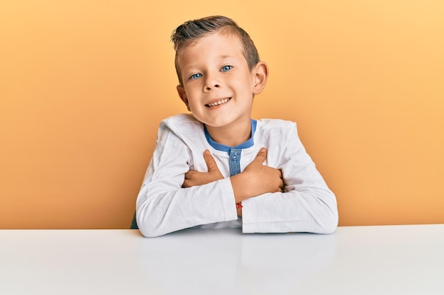 Adorable caucasian kid wearing casual clothes sitting on the table happy face smiling with crossed arms looking at the camera positive person