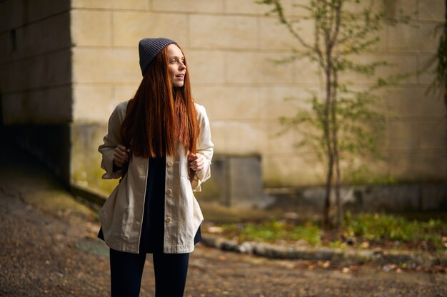 Adorable caucasian redhead female in coat and hat traveler with backpack walking in historical and a...