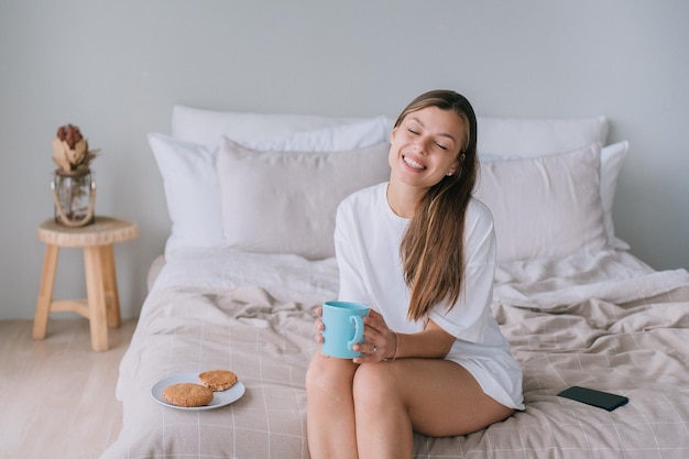 Adorable caucasian young woman sitting on bed holds cup of coffee eyes closed smiles Close up of beautiful Italian girl enjoys sunny morning at home Happy people Domestic leisure Cozy home