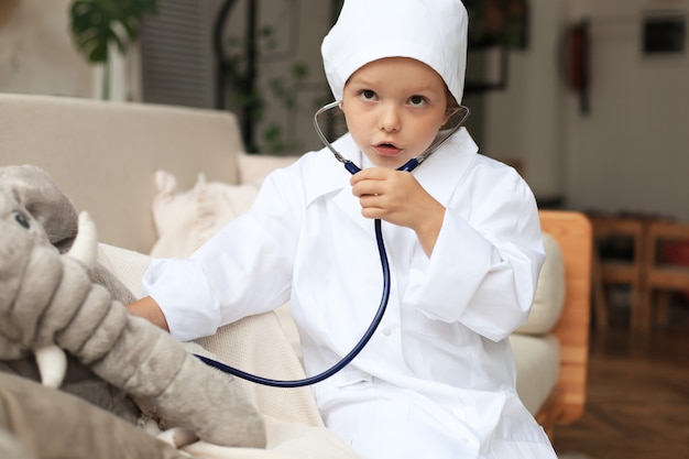 Adorable child dressed as doctor playing with toy elephant, checking its breath with stethoscope.