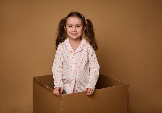 Adorable child European 4 years old baby girl with two wavy ponytails in a bright pajamas looking at camera standing inside a cardboard box isolated over beige background with copy space for ads