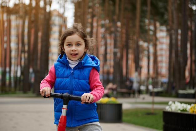 Adorable child girl riding a kick scooter in the city park on a sunny day in early autumn or spring Healthy and active lifestyle leisure activity