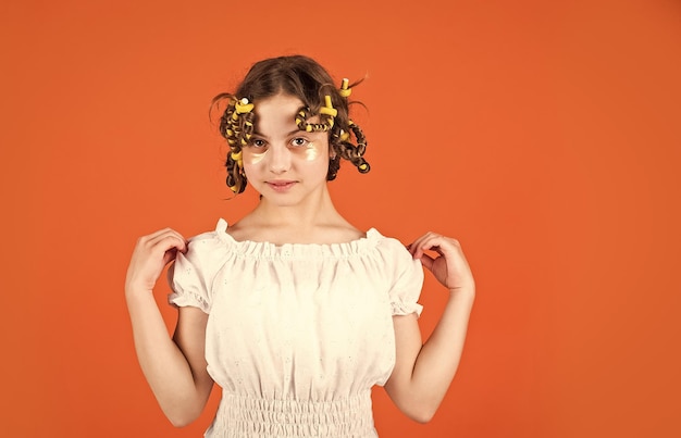Фото Очаровательная детская прическа дочь с бигуди на голове смеется парикмахерская салон женской красоты советы по укладке подростковые хобби маленькая девочка керлинг волосы используя бигуди оранжевый фон