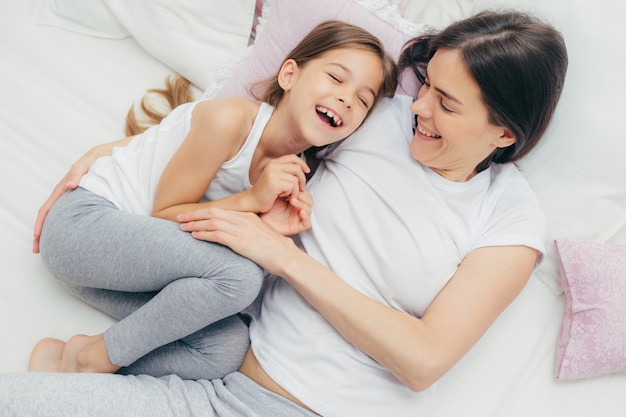 Adorable child and her mother have fun together in bed, tickle each other, smile joyfully, play after good sleep