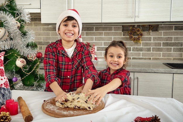사랑스러운 아이들은 집에 있는 부엌에서 크리스마스 빵을 위해 달콤한 반죽을 반죽하고 굴리고, 달콤하게 웃고, 카메라를 바라보고 있습니다