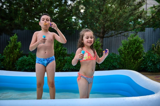 Adorable children siblings blowing soap bubbles while having fun in the inflatable water pool on a hot summer day