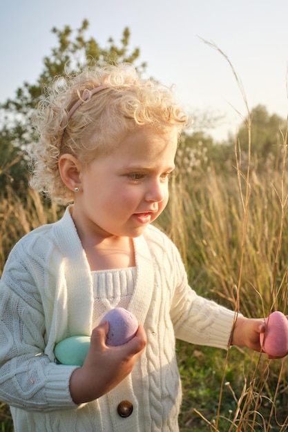 Adorable content girl in white sweater collecting colorful Easter eggs on grassy meadow in sunny spring nature