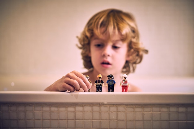 Adorable curious little boy with curly blond hair sitting in bathtub and playing with mini figures of various professions at home
