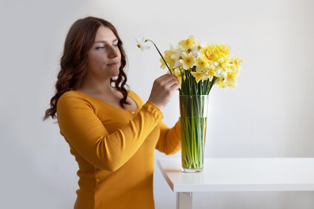 Adorable curly hair girl puts a transparent vase with spring flowers on a white table in the room. Housewife taking care of coziness and decor.