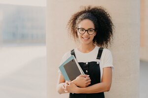 照片可爱的卷发女学生穿着白色休闲t恤和工作服，拿着笔记本或教科书