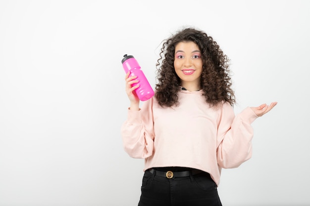 白の上に立っているピンクの水のボトルを持つ愛らしい巻き毛の女性。