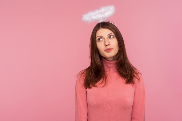 Adorable cute brunette woman in pink sweater standing with halo over head angelic innocent expression faith Indoor studio shot isolated on pink background