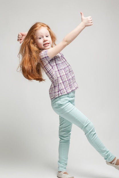 Adorable cute redhead little girl in plaid shirt, bright blue trousers and white boots