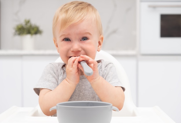 Adorable cute sad caucasian blonde baby girl crying eating from silicone plate at high chair in kitchen, with spoon in mouth, looking at camera.Healthy food for kid concept.