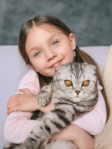 Photo adorable cute smiling girl hugging scottish gray cat on the sofa at home