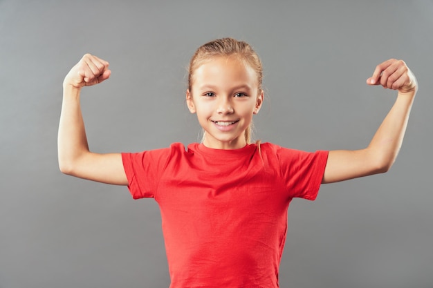 Adorabile bambina in camicia rossa che mostra i muscoli del braccio e sorride mentre sta in piedi su uno sfondo grigio