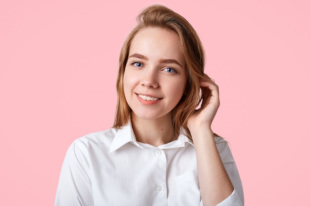 adorable female college student with blue eyes, healthy pure skin, wears white shirt, poses on pink, has gentle pleasant smile. People, positive emotions concept.