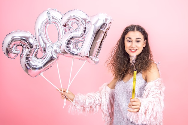 Adorable festively dressed brunette girl with curly hair on a pink  wall looks at an extinguished fireworks candle, with silver balloons for the new year concept