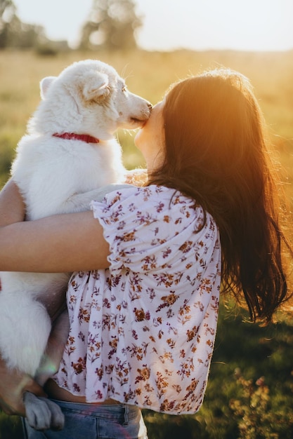 Foto adorabile cucciolo soffice che bacia ragazza bellissimo momento atmosferico elegante giovane donna che abbraccia un simpatico cucciolo bianco nella calda luce del tramonto nel prato estivo concetto di adozione