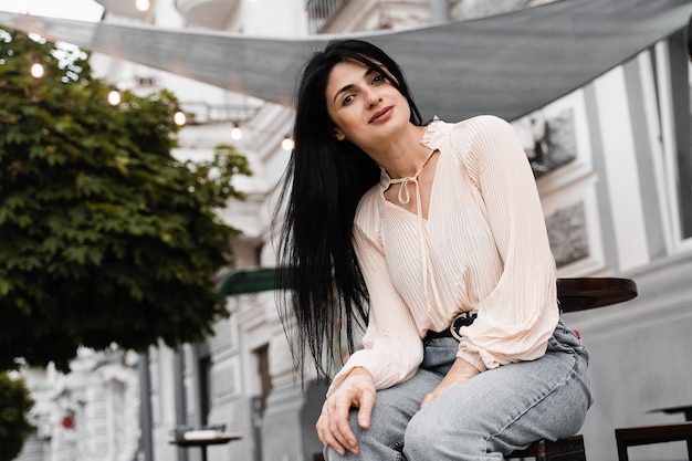 Adorable Georgian woman is sitting in cafe outdoor and posing Fashion girl with different emotions waiting her order on summer terrace