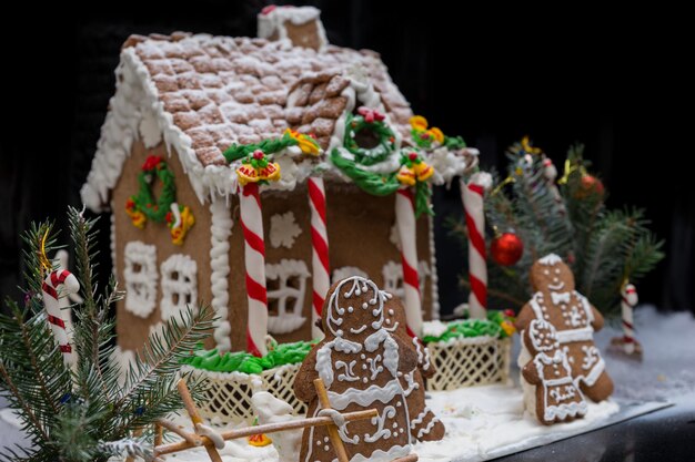 Famiglia adorabile del pan di zenzero vicino alla casa di pan di zenzero fatta in casa coperta di neve con un rametto di albero di natale su sfondo scuro. mockup per offerte stagionali e cartolina delle vacanze