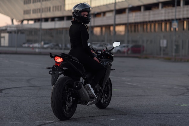 검은 꽉 몸 슈트와 전면 헬멧에 사랑스러운 소녀는 저녁에 도시 야외 주차장에서 세련된 오토바이 타기. 자유와 활동적인 라이프 스타일 개념. 완전히 검은 색.