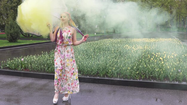 Adorable girl in a dress with a floral print with rainbow braids and makeup. Dances with an umbrella hiding in yellow smoke against the backdrop of flower bed with tulips