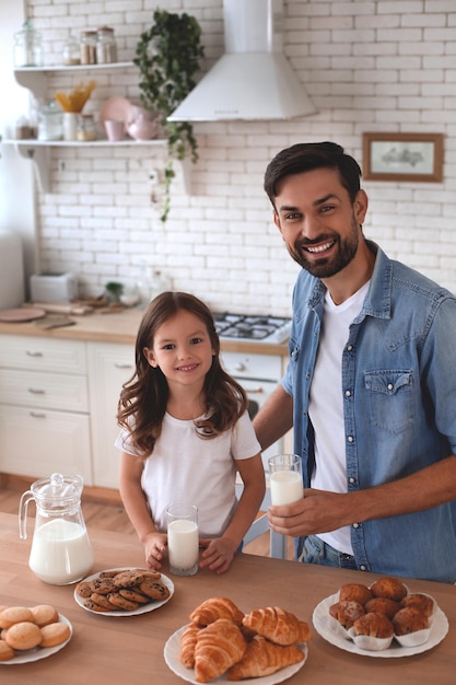 カメラを見ているテーブルにミルクとクッキーのグラスを保持している愛らしい女の子と彼女の父親