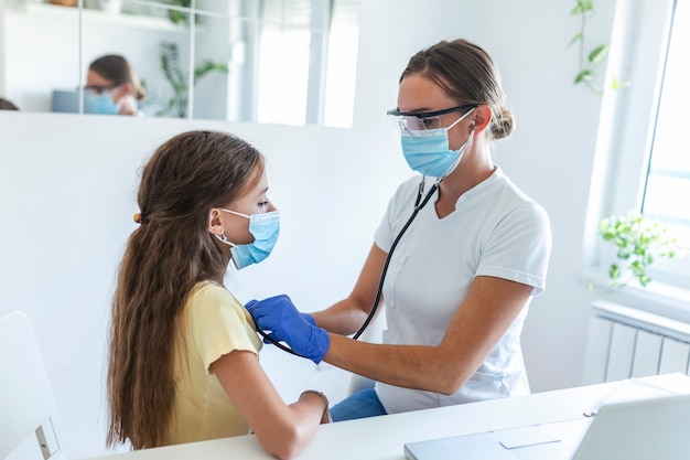 Adorable girl is sitting at her doctor's office. her doctor is
using a stethoscope to listen to her chest. both her and her doctor
are wearing a face mask to prevent the spread of germs.