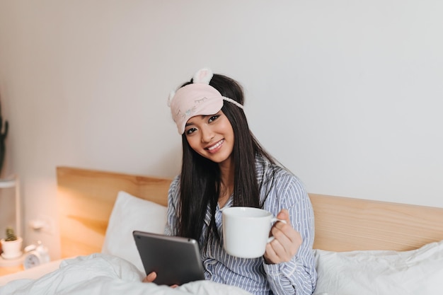 Adorable girl in striped pajamas posing with big cup and tablet Woman in sleep mask is lying in white bed