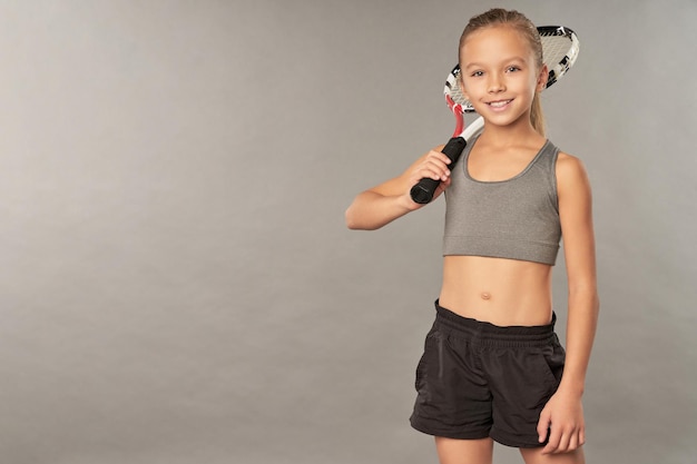 Adorable girl with tennis racket standing against gray background