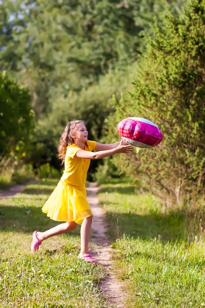 노란 드레스를 입은 사랑스러운 소녀가 꽃 모양의 축제 생일 다채로운 풍선을 들고 있습니다. 야외에서 행복한 아이