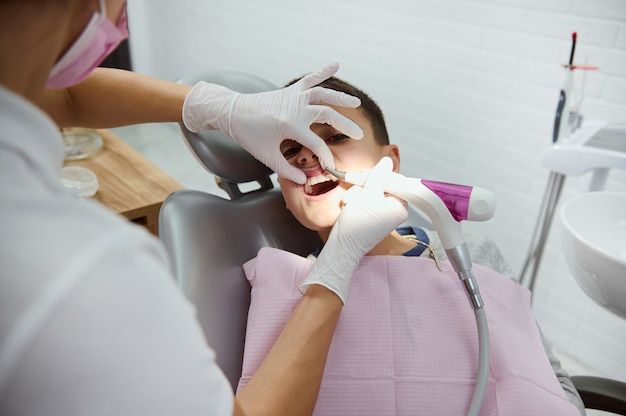 Adorable handsome child, school boy sitting in dentist's chair during paediatric dentist visit in modern white dental clinic, receiving medical oral cavity treatment. Dental oral hygiene concept