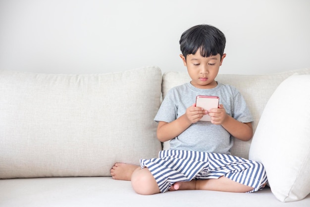 An adorable happy Asian boy wearing a gray shirt and bluewhite striped shorts is having fun playing with his smartphone on a cream sofa looking at the mobile screen