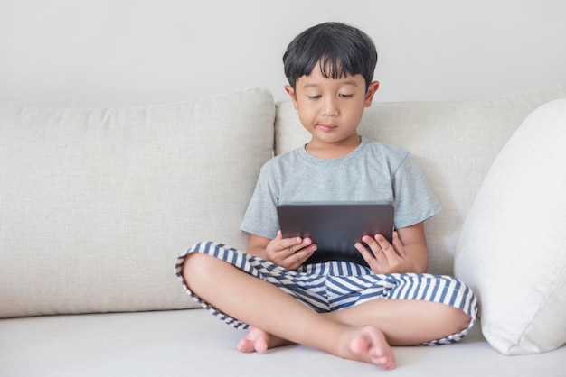 Adorable happy Asian boy wearing a gray shirt and bluewhite striped shorts is having fun playing with his tablet on a cream sofa looking at the mobile screen